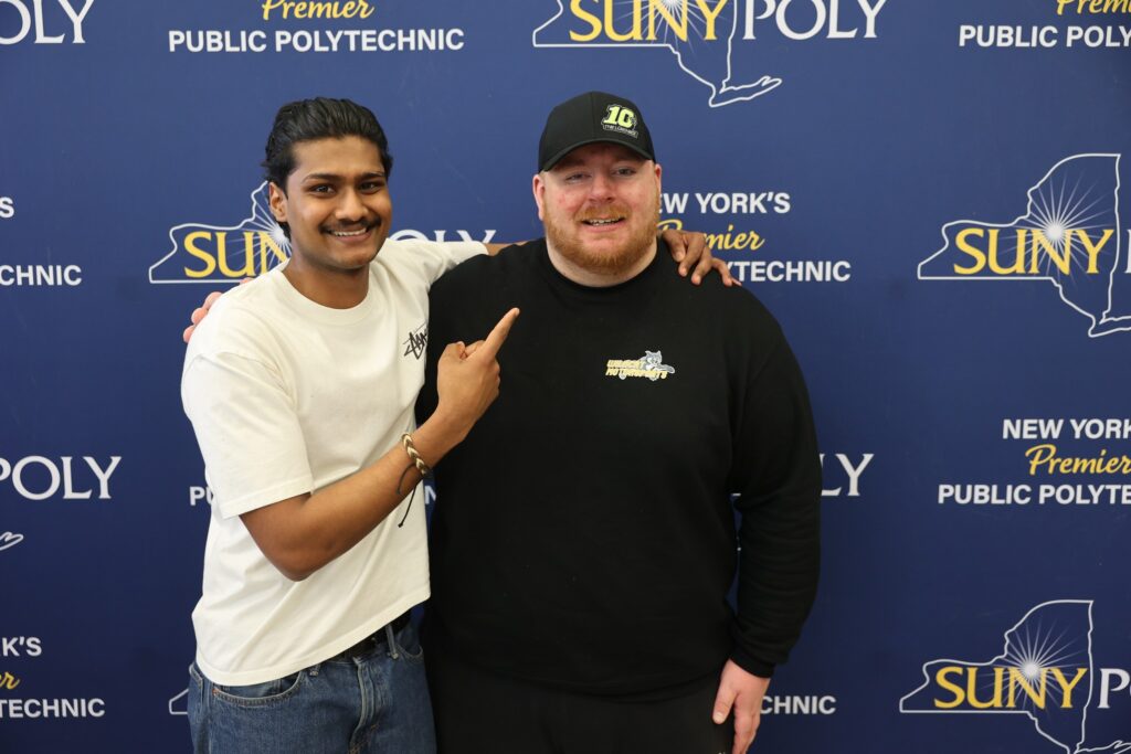 Two students posing together in front of a SUNY Poly backdrop.
