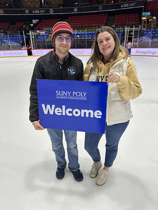 Two people holding the SUNY Poly Alumni Association Welcome sign on center ice.