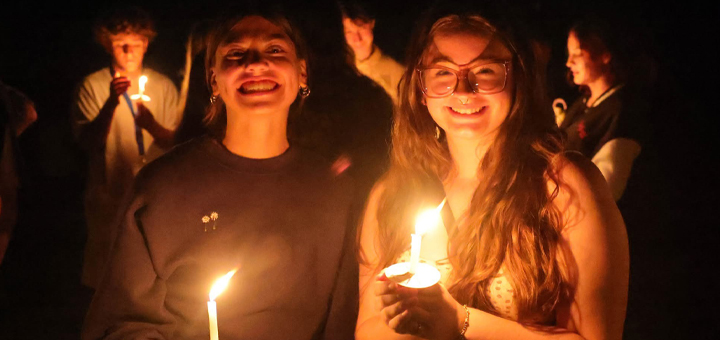 Two students smiling and holding lit candles during a nighttime campus vigil with others gathered in the background.
